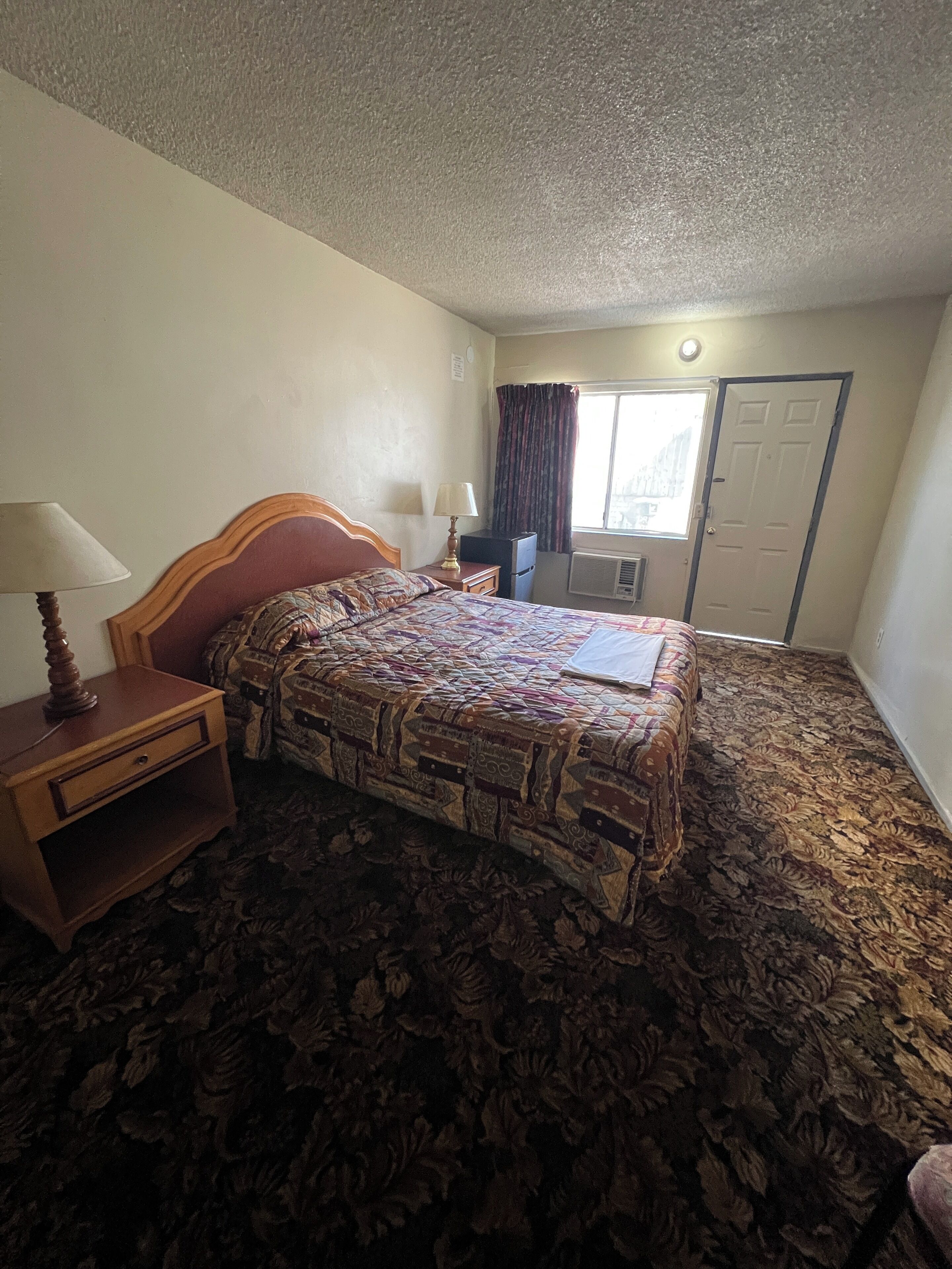 A hotel room with a large bed, a nightstand with a lamp, a dresser, and a window with curtains.