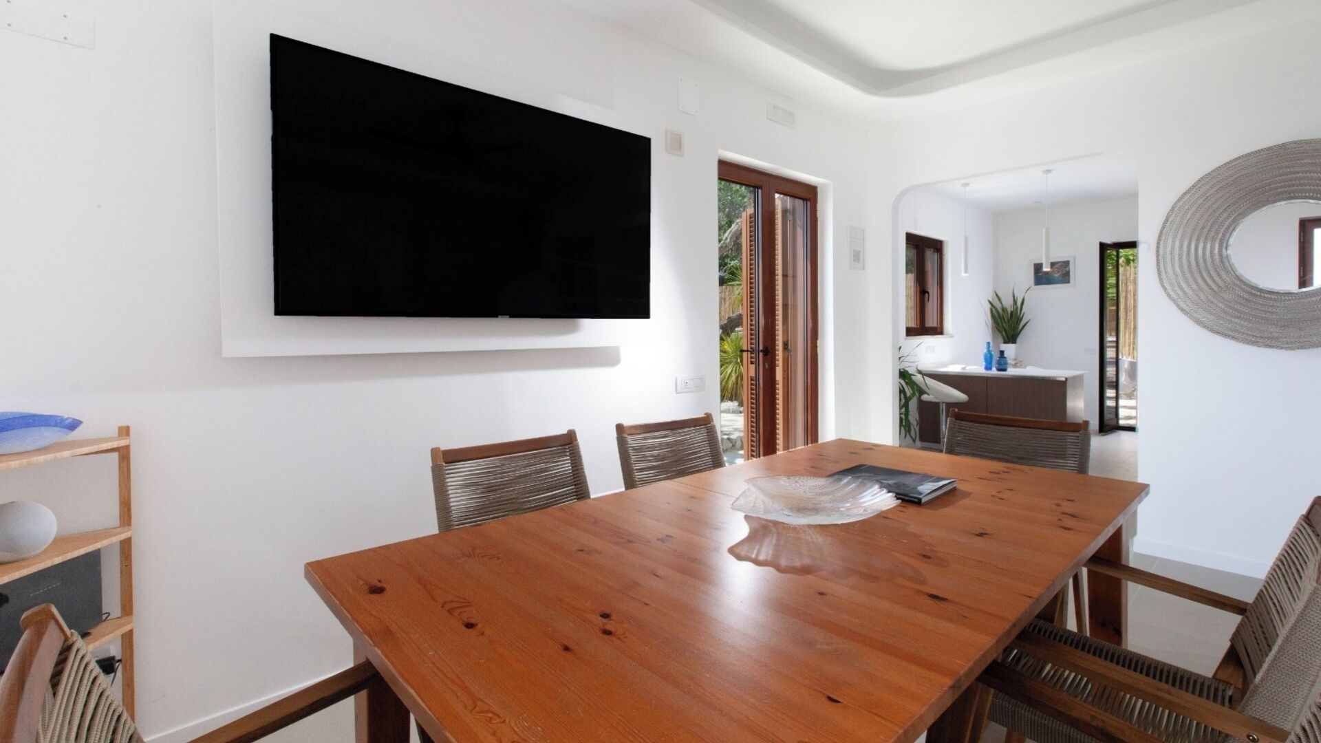 A dining area with a wooden table, chairs, a flat-screen TV mounted on the wall, and a view of the outdoors through a glass door.