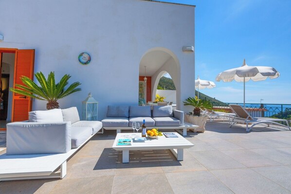 Ein moderner Sitzbereich im Freien mit weißen Möbeln, einem Tisch mit Früchten und Blick auf das Meer.