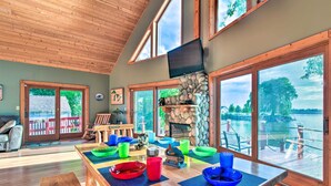 A spacious living room with a wooden ceiling, large windows, a stone fireplace, and a dining table with colorful bowls.
