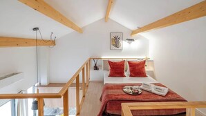 A high-ceilinged room with exposed wooden beams, a bed with red bedding, a bedside table with a tray, and a framed artwork on the wall.