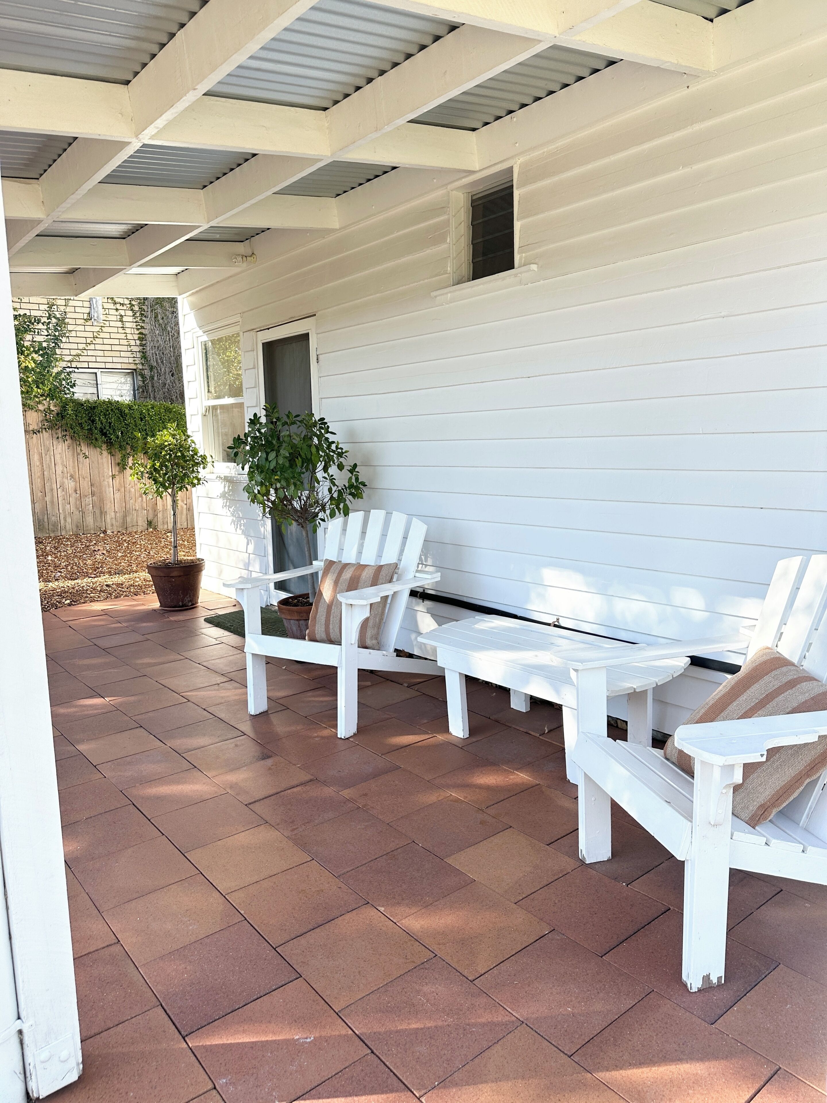 Un patio cubierto con sillas Adirondack blancas y una mesa, una planta en maceta y una valla de madera al fondo.