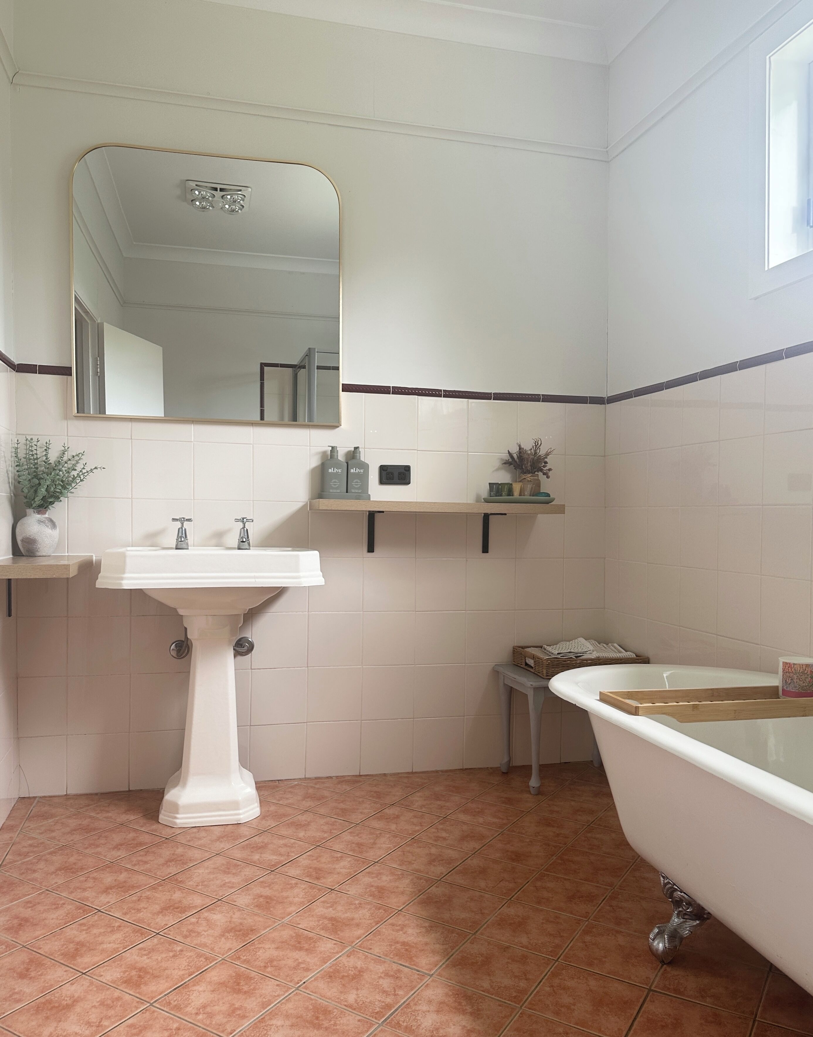A bathroom with a pedestal sink, a large mirror, a bathtub, and a small shelf with toiletries.