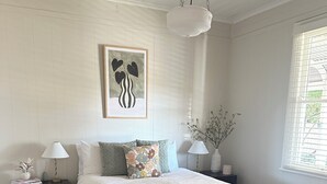 Ein Schlafzimmer mit einem Bett, Nachttischen, einer Pendelleuchte, einem gerahmten Kunstwerk und einem Fenster mit Jalousien.