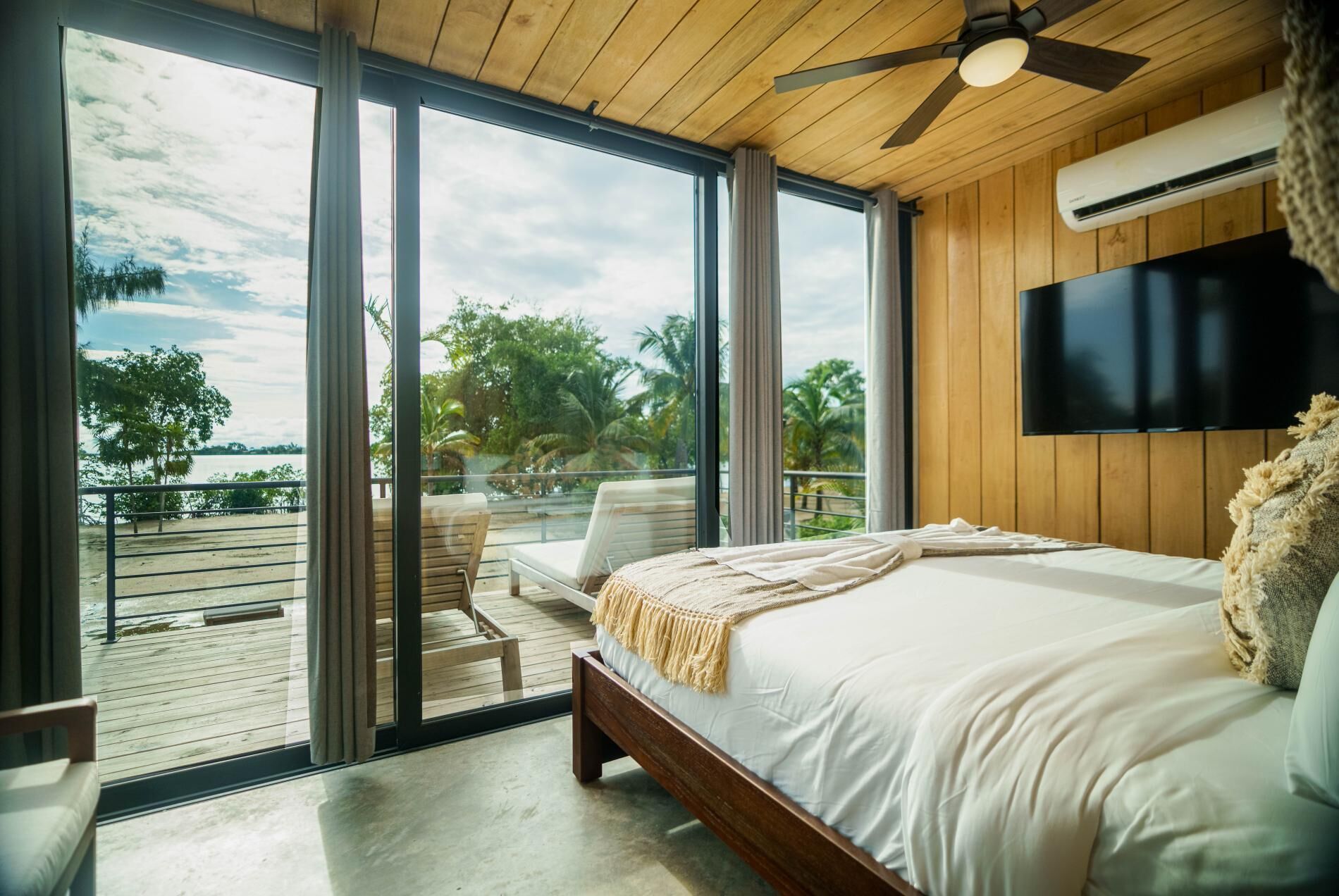 A bedroom with a large bed, a ceiling fan, a TV, and a view of the outdoors.