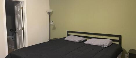 A bedroom with a bed, a nightstand, a lamp, and a door leading to a bathroom.