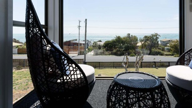 A room with a large window overlooking a coastal view, featuring a black woven chair and a small round table. - Ocean View Retreat at Greens Beach (Greens Beach)