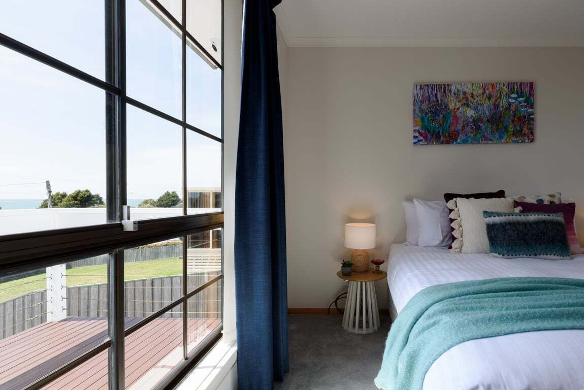 A hotel room with a large window, a bed with multiple pillows, a small bedside table with a lamp, and a colorful abstract painting on the wall.