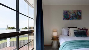 A hotel room with a large window, a bed with multiple pillows, a small bedside table with a lamp, and a colorful abstract painting on the wall. - Ocean View Retreat at Greens Beach (Greens Beach)
