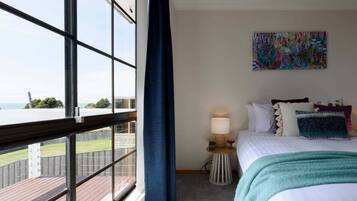 A hotel room with a large window, a bed with multiple pillows, a small bedside table with a lamp, and a colorful abstract painting on the wall.