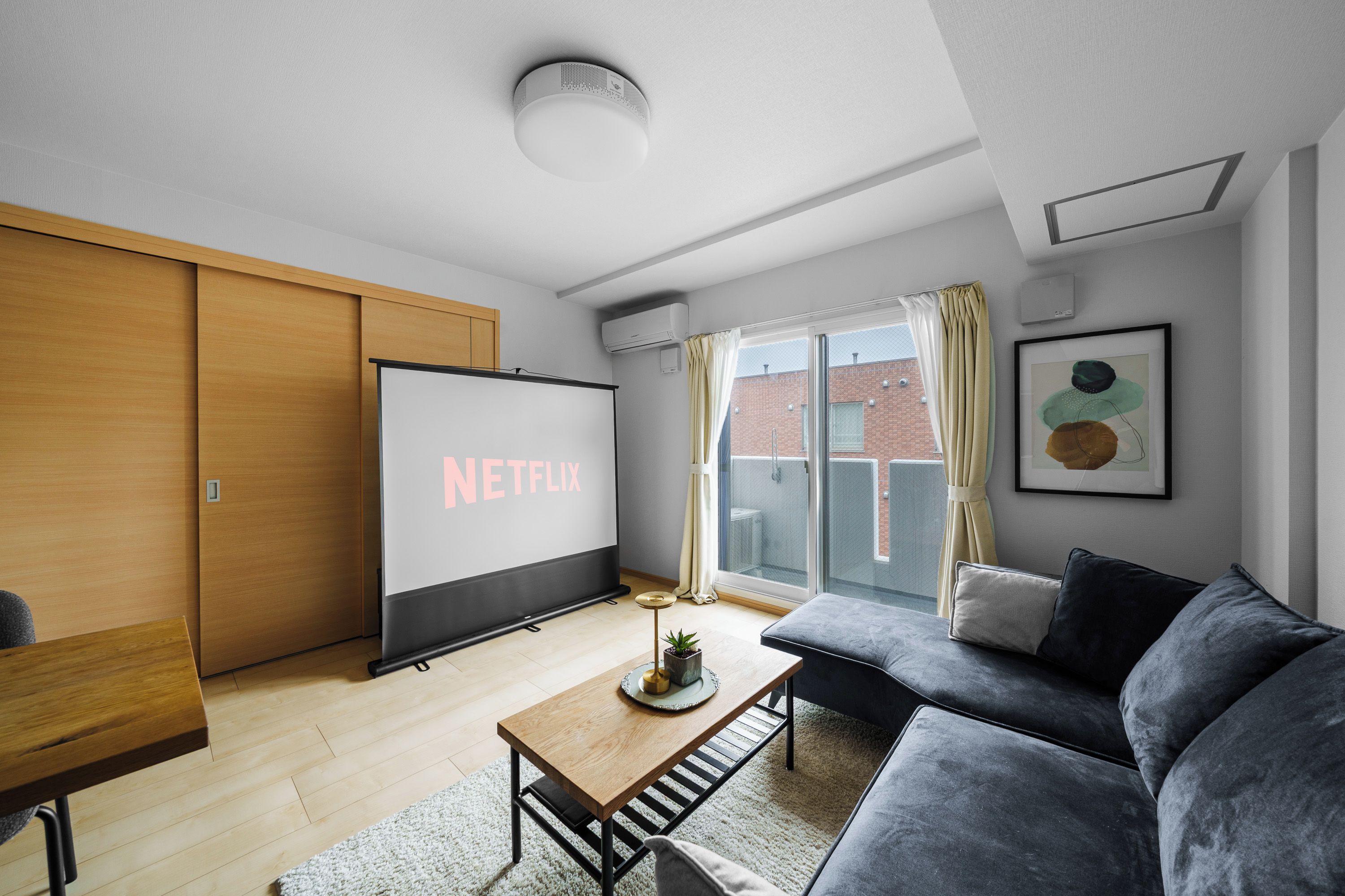 A modern living room with a large flat-screen TV displaying 'NETFLIX,' a wooden table, a grey sofa, and a view of a brick building through a large window.