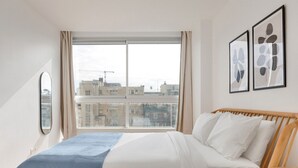Une chambre moderne avec une grande fenêtre, un lit recouvert de linge de lit blanc et deux œuvres d’art abstraites encadrées accrochées au mur.