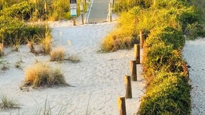 Un sentier de sable qui longe la plage et mène à l’océan, bordé de végétation et d’une clôture en bois.