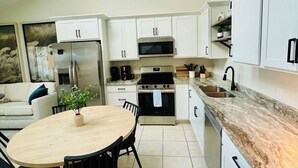 Une cuisine moderne avec des meubles blancs, un réfrigérateur en acier inoxydable, un four à micro-ondes et une cuisinière. On y trouve une table à manger avec des chaises et un luminaire suspendu.