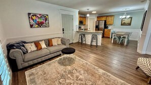 A living room with a sofa, ottoman, and rug, a kitchen with bar stools, and a dining area with a table and chairs. - The Grove Getaway-Centrally Located (Pleasant Grove)