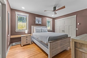 A bedroom with a wooden bed, a nightstand, a desk, a ceiling fan, and a window with a view of trees. - Peaceful Forest Escape by Lake Elmore (Elmore)