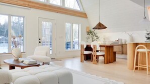 Un salon spacieux avec une grande fenêtre, un coin repas avec des meubles en bois et une cuisine dotée d’un plan de travail en bois.