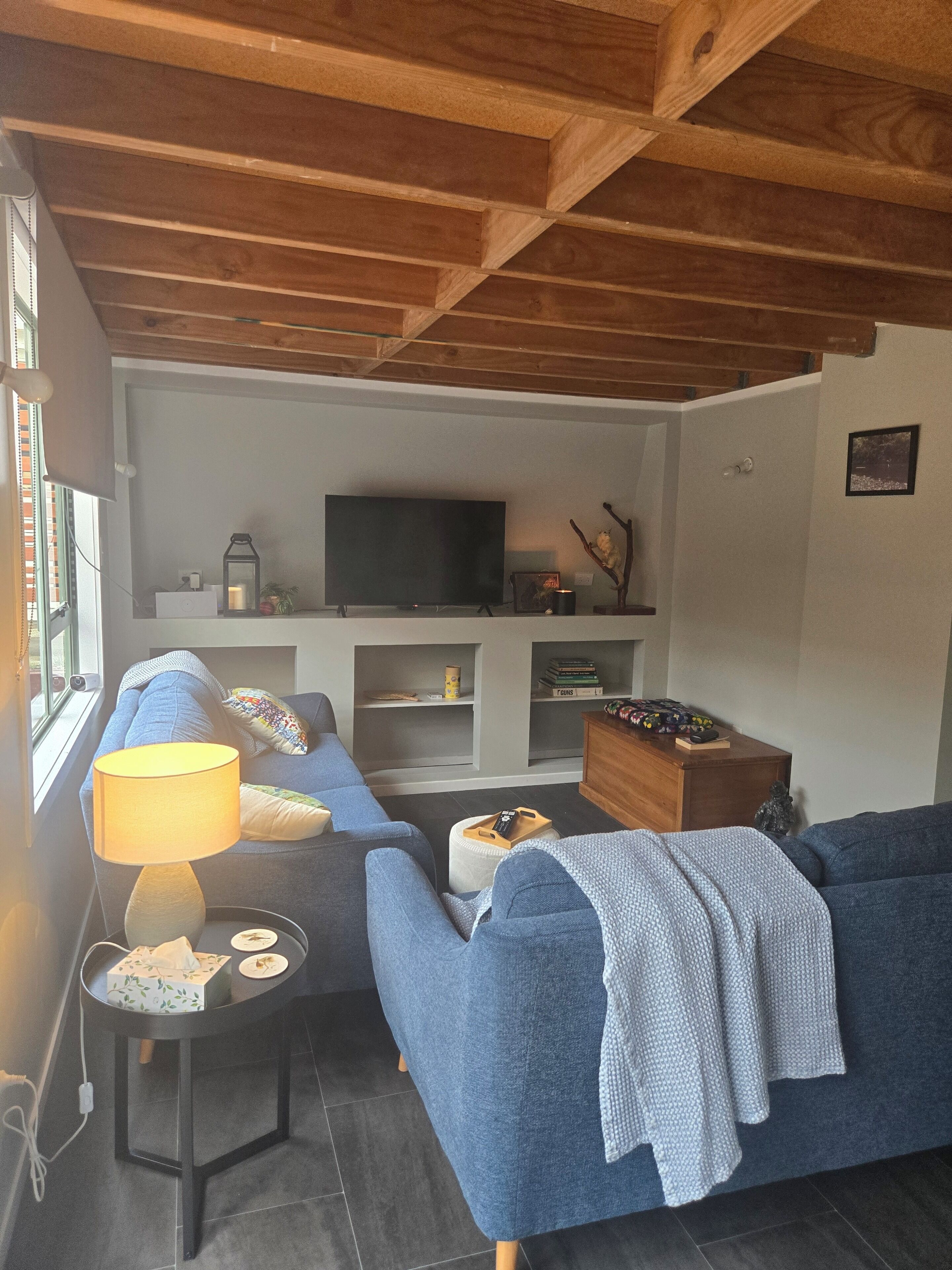 A cozy living room with a blue sofa, a small side table with a lamp, a television on a white stand, and a wooden cabinet.