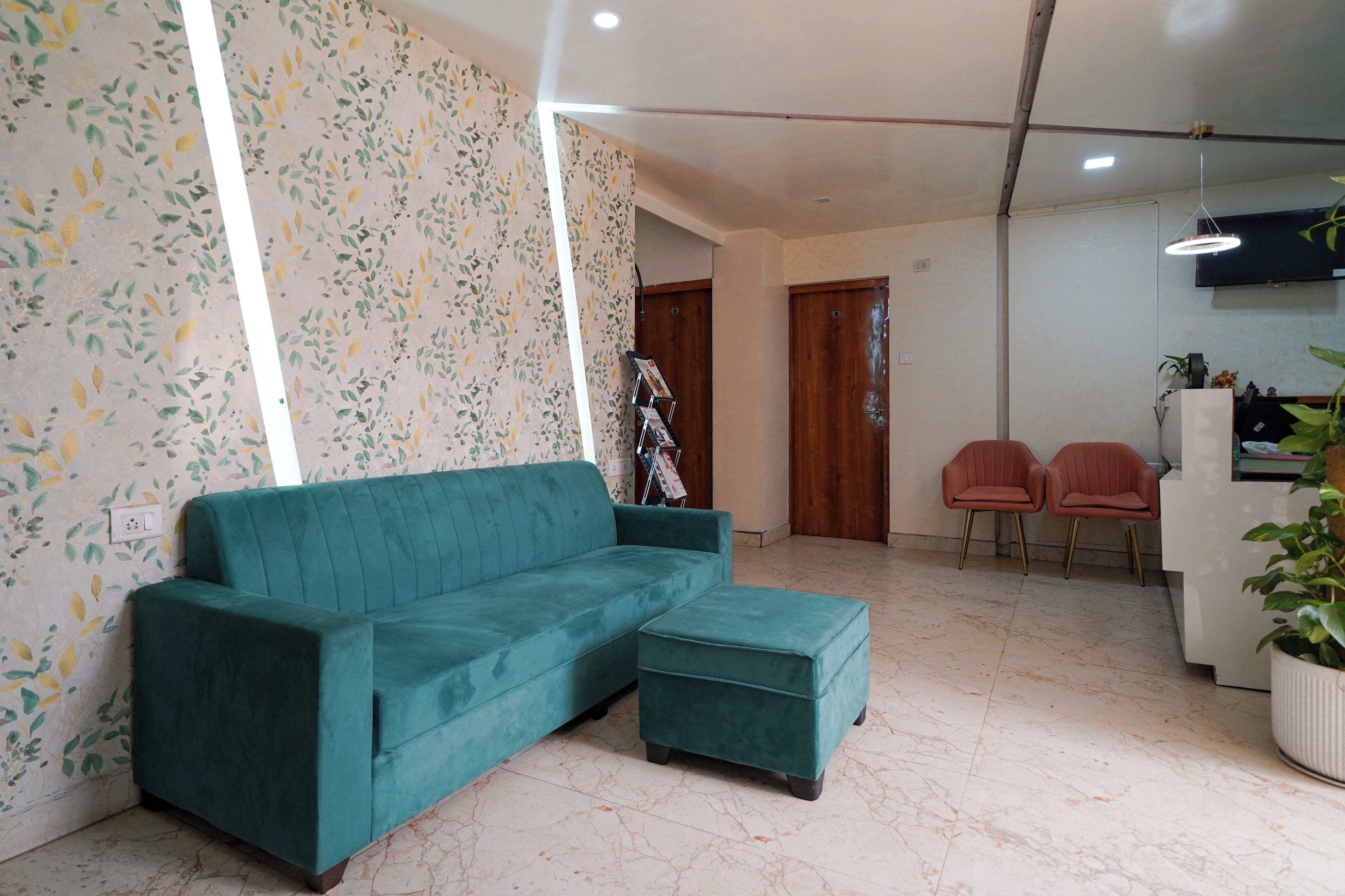 A living room with a teal sofa, a matching ottoman, a wooden door, a small table with two chairs, and a TV mounted on the wall.