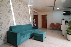 A living room with a teal sofa, a matching ottoman, a wooden door, a small table with two chairs, and a TV mounted on the wall. - VH Hotels Domlur (Bengaluru)