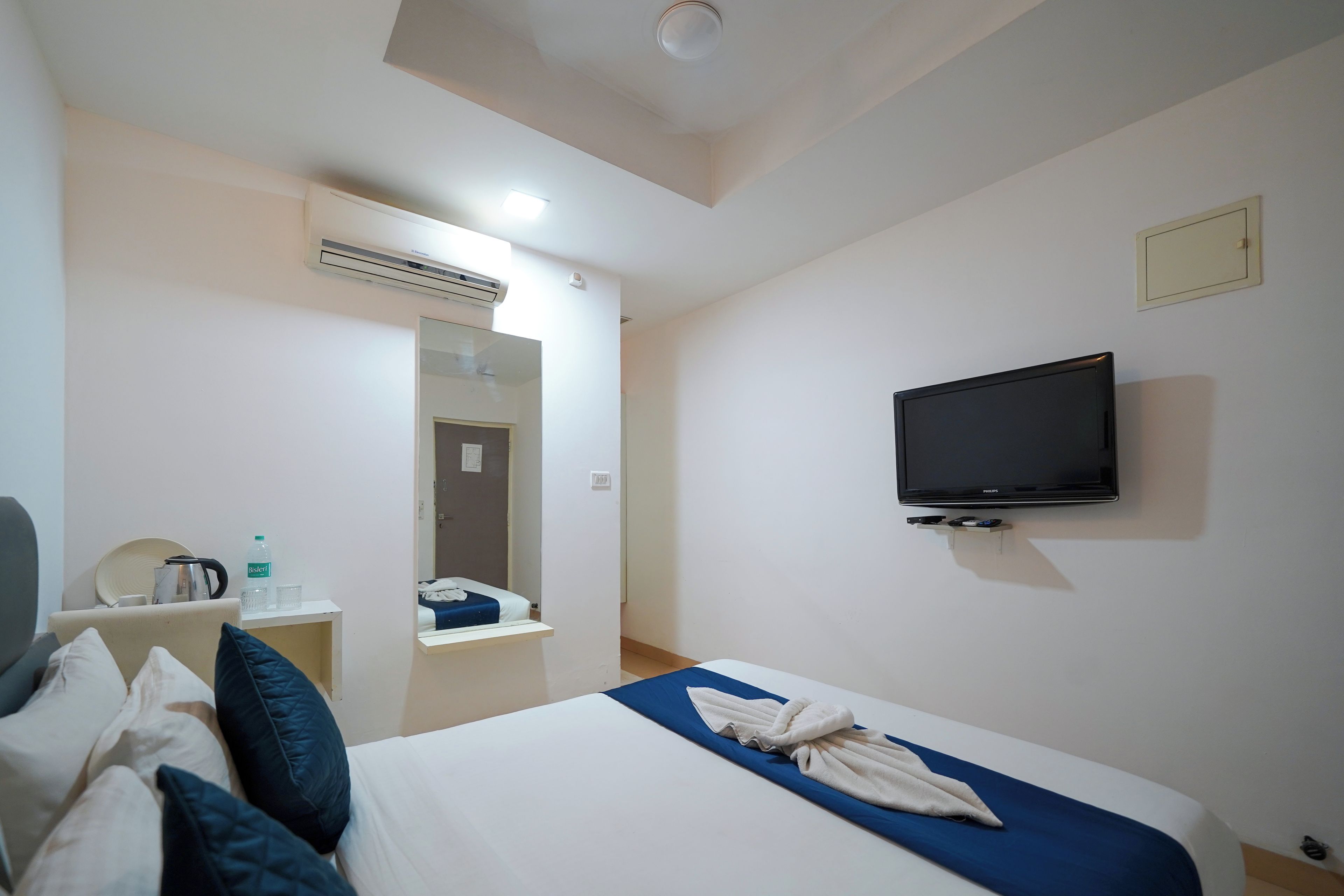 A hotel room with a bed, a television, a mirror, and a small table with a kettle and water bottle.