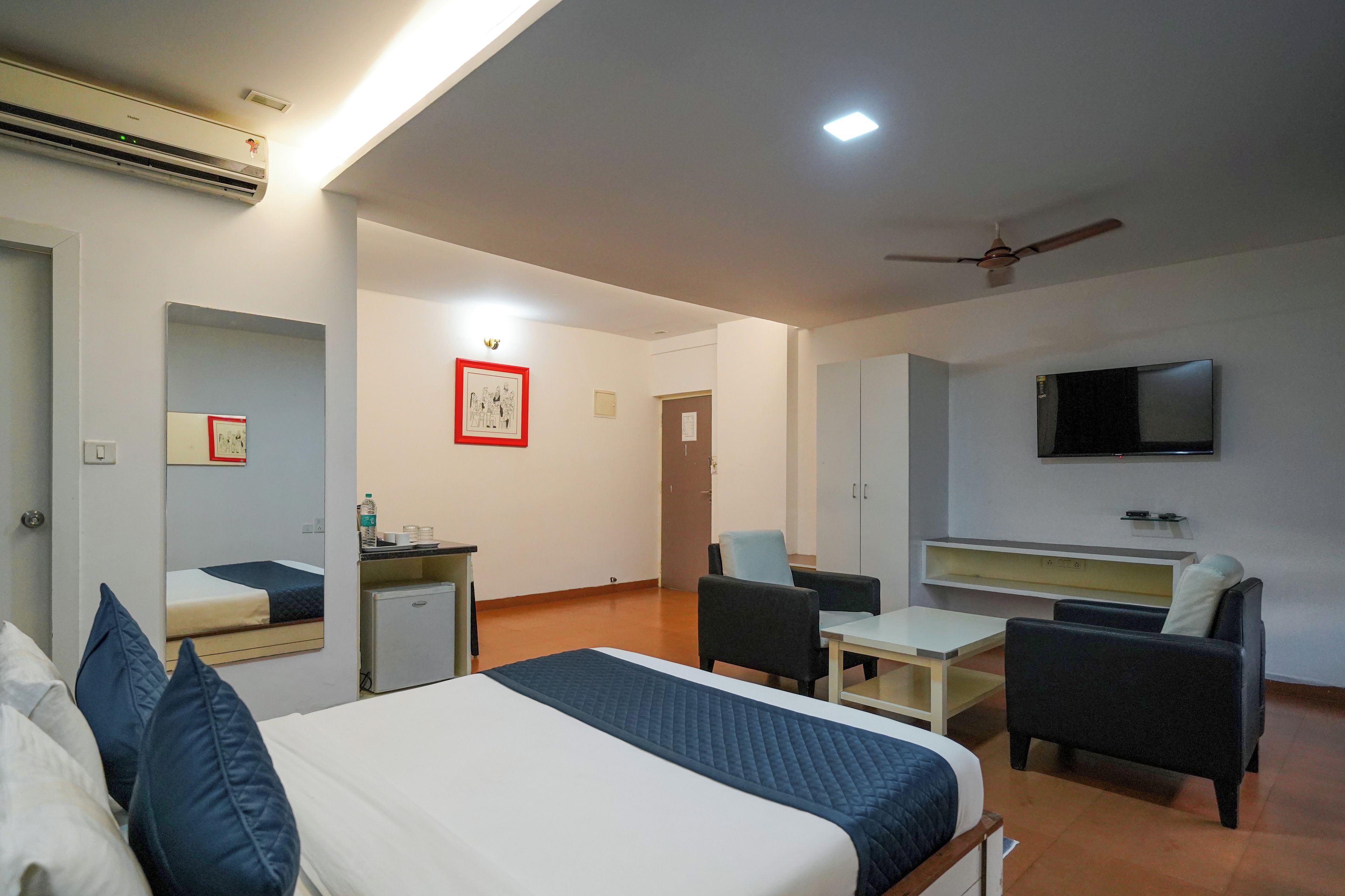 A hotel room with a bed, desk, chairs, a television, and a ceiling fan.