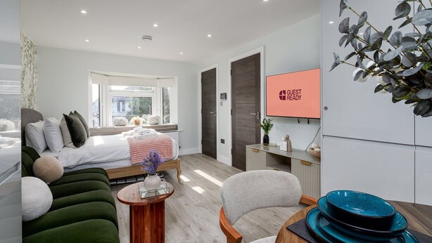 Een moderne hotelkamer met een bed, een groene bank, een houten bijzettafeltje en een televisie waarop staat: 'GUEST READY'.