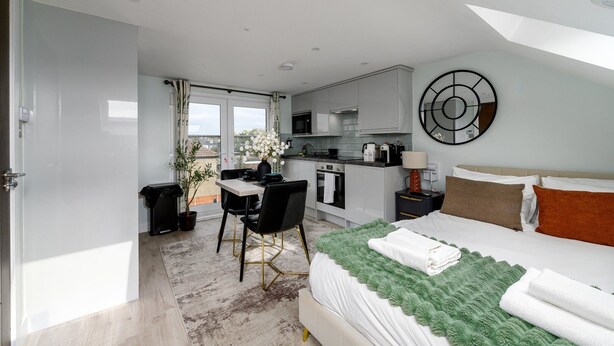 Een moderne hotelkamer met een bed, een groene plaid, een zithoek met een tafel en stoelen, een keuken, en een balkon met uitzicht.