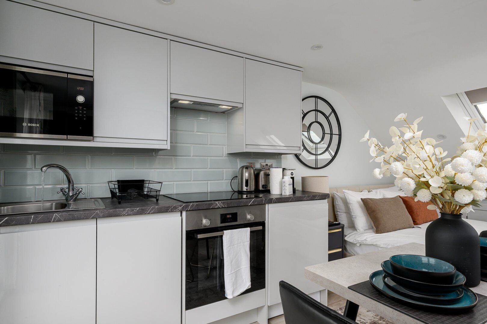 A modern kitchen with white cabinets, a Bosch microwave, and a sink. There's a dining area with a table, chairs, and a vase with white flowers.