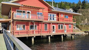 A two-story waterfront house with a balcony, wooden deck, and a dock extending into the water.