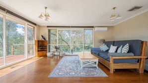 A spacious living room with a large window, a wooden sofa, a coffee table, and a bookshelf. - Tranquil Cottage with Valley & Hills Views (Wonga Park)