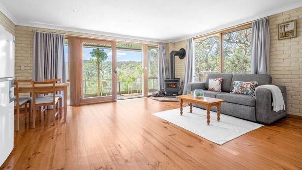 A spacious living room with a fireplace, a sofa, a dining table with chairs, and large windows offering a view of the outdoors. - Tranquil Cottage with Valley & Hills Views (Wonga Park)