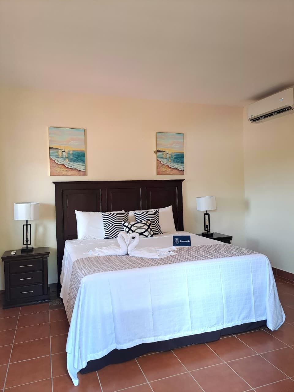Une chambre d’hôtel comprenant un lit, deux tables de chevet, une petite table, un climatiseur fixé au mur et deux tableaux.