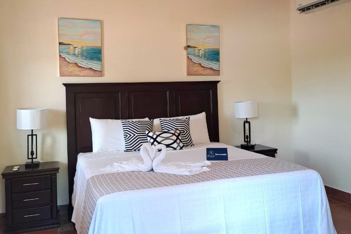 A hotel room with a bed, two bedside tables, a nightstand, a wall-mounted air conditioner, and two paintings.