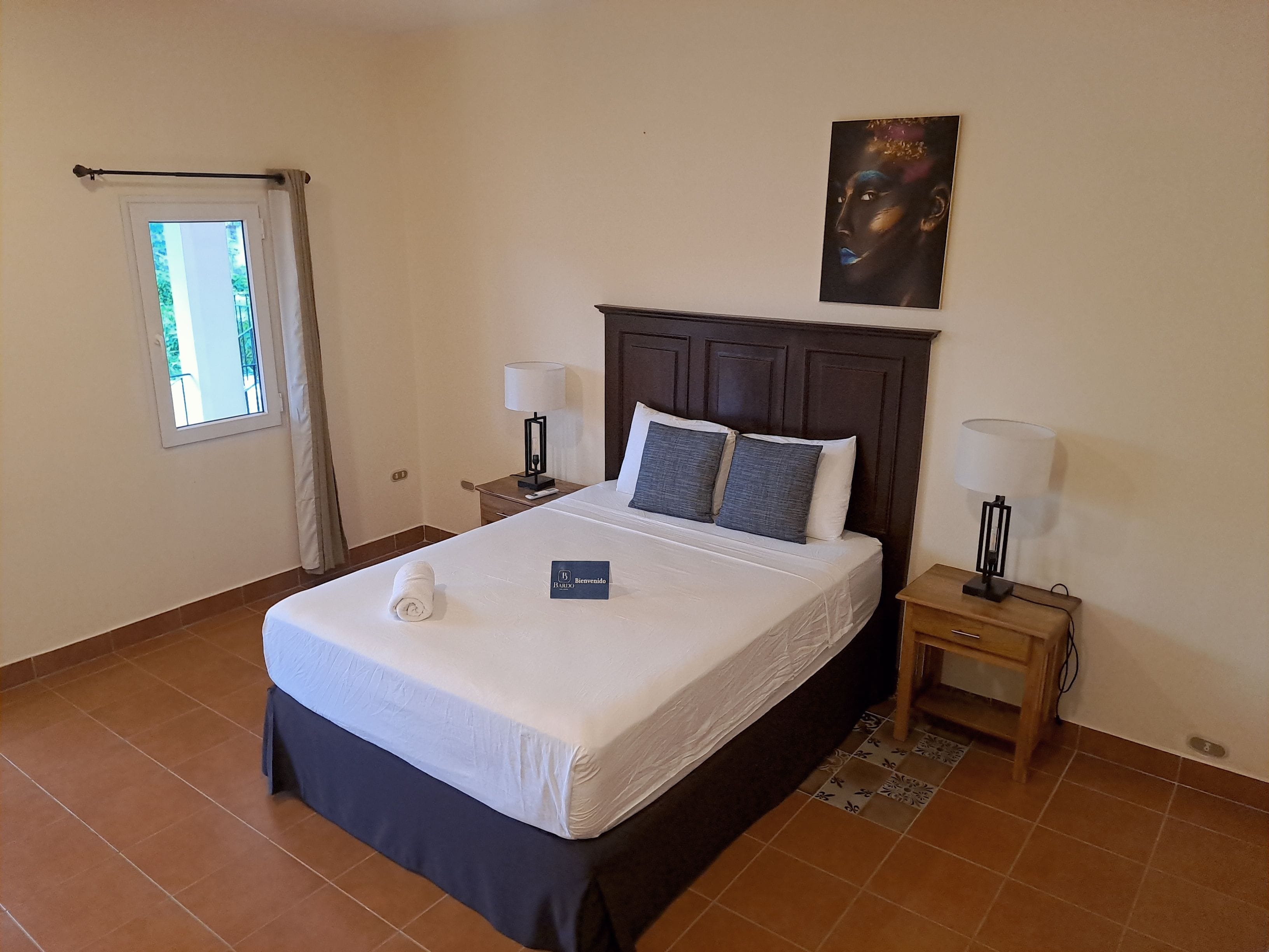 A hotel room with a bed, two bedside tables with lamps, a window with curtains, a wall-mounted painting, and a welcome sign.
