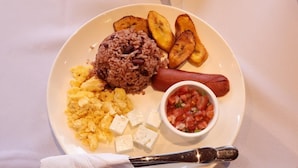 A plate with scrambled eggs, black beans rice, a hot dog, fried plantains, and a small bowl of salsa.