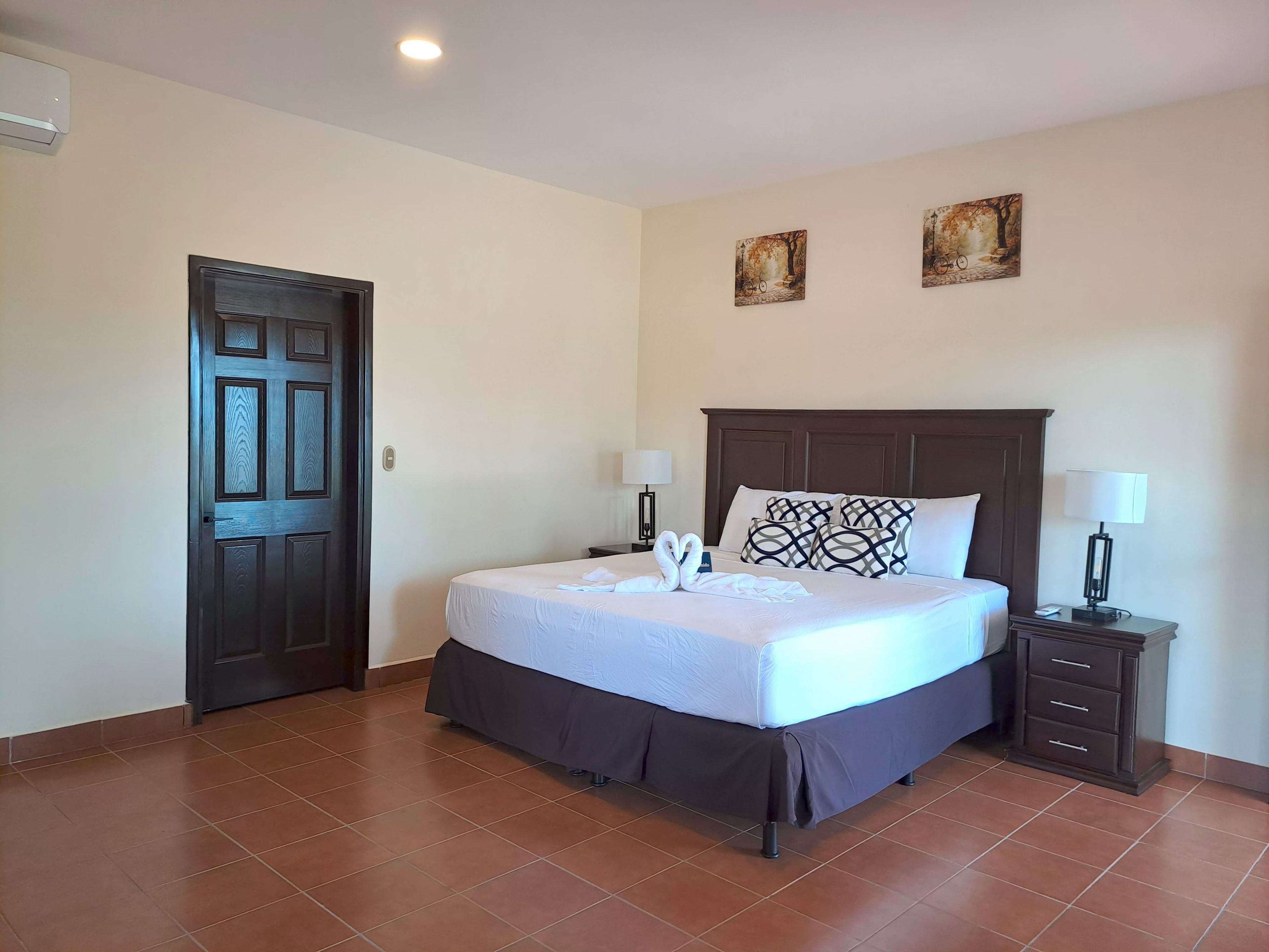 A hotel room with a bed, two wall-mounted paintings, a nightstand with a lamp, and a door.