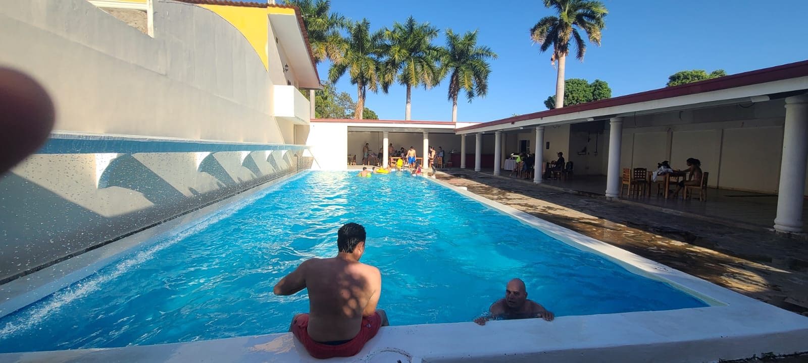 A swimming pool with people in it, surrounded by a covered area with tables and chairs.