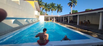 Una piscina con gente, rodeada por un área techada con mesas y sillas.