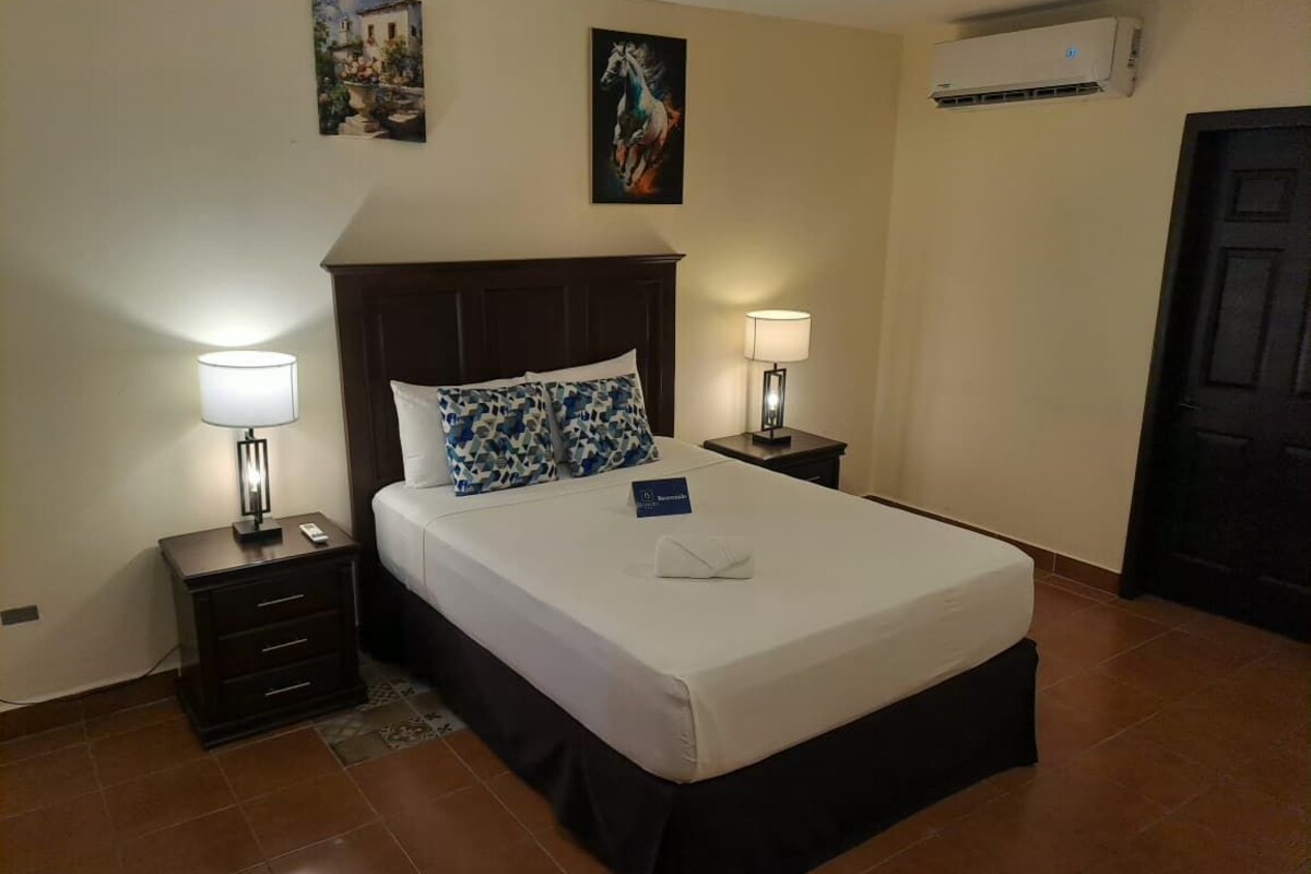 A hotel room with a bed, two bedside tables with lamps, a nightstand, a wall-mounted air conditioner, and a door.