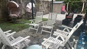 Mobiliario exterior con sillas blancas y una mesa de vidrio, rodeado de un patio de hormigón y una zona ajardinada.