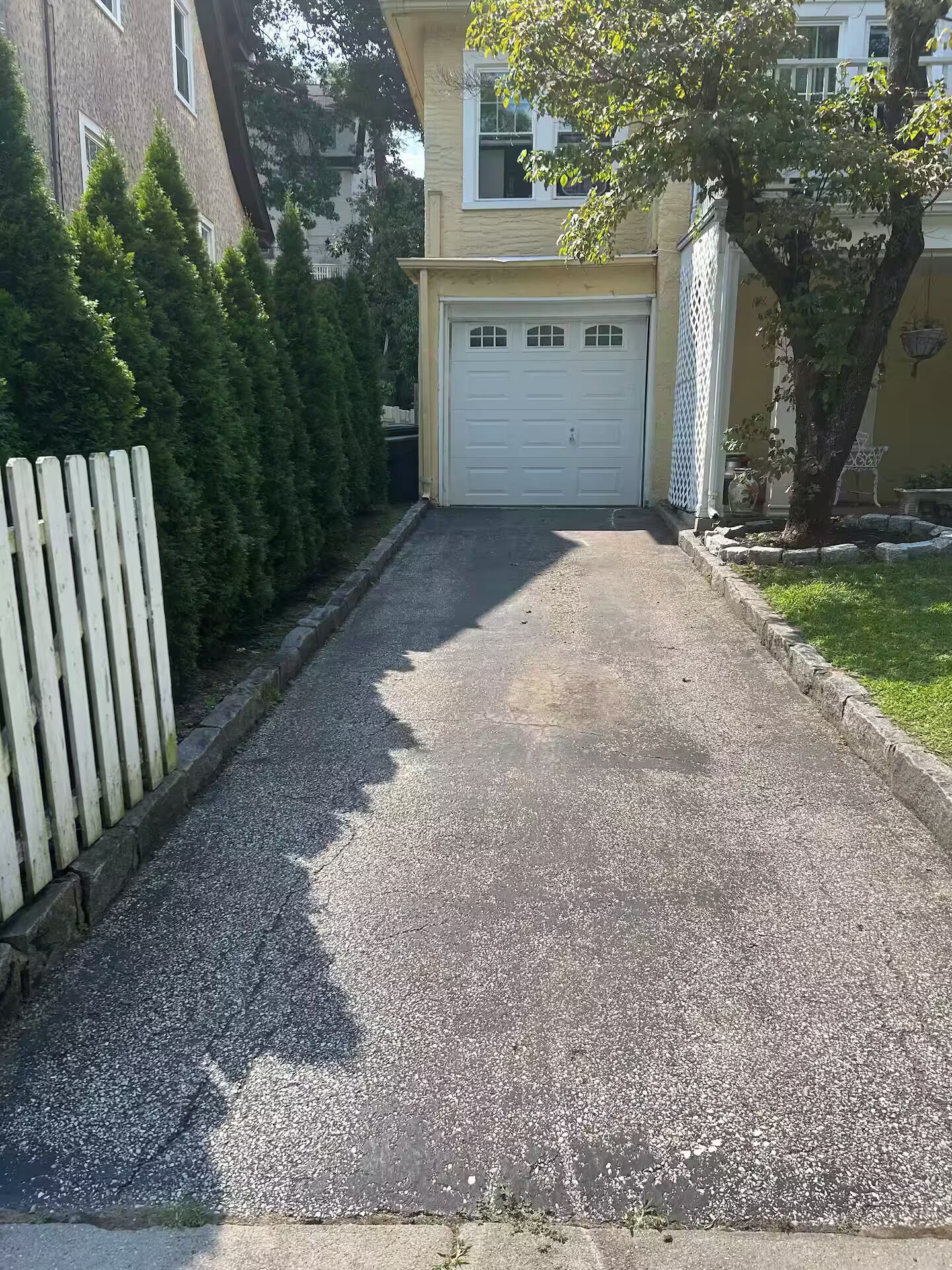 En uppfart som leder till en garageport, med en trästaket på ena sidan och en rad vintergröna träd på den andra.
