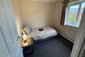 A small, single-bed room with a window, a bedside table with a lamp, and a radiator.