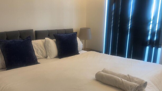 A neatly made bed with a folded towel, a lamp, and a window with vertical blinds. - London 1 Bed Apartment close to Barking Station (Barking)