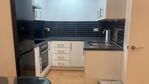 A compact kitchen with white cabinets, a black countertop, and a tiled backsplash. - London 1 Bed Apartment close to Barking Station (Barking)