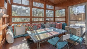 A cozy wooden cabin with a large windowed porch, a cushioned bench, and a metal table.