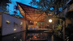 Un área de comedor al aire libre con un espacio de descanso cubierto, una piscina y vegetación circundante.