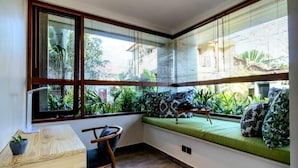Habitación de esquina con una mesa de madera, una silla, un banco acolchado con cojines estampados y vistas a zonas verdes a través de ventanas grandes con persianas.