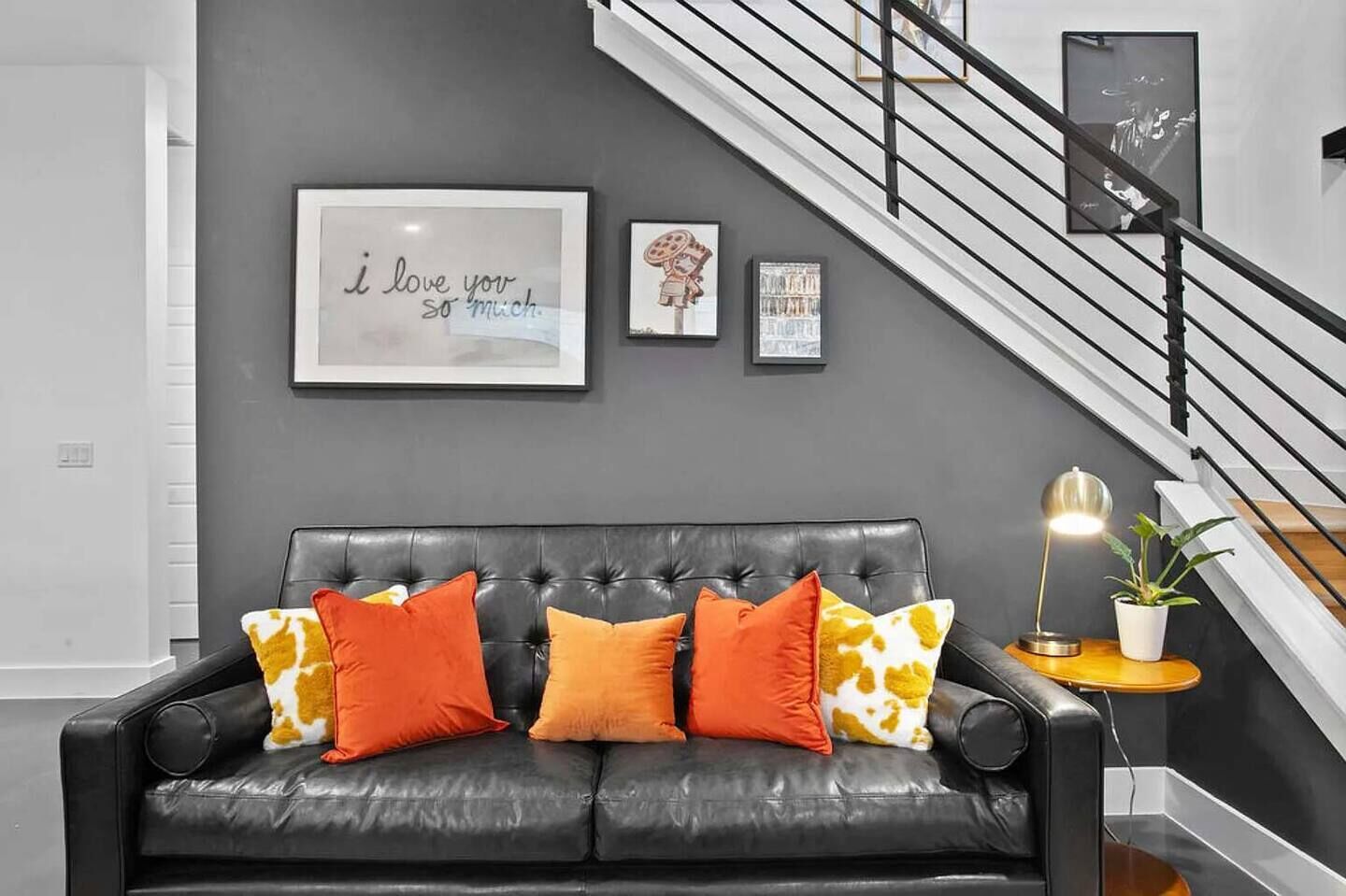 Una sala de estar moderna con un sofá de cuero negro, cojines estampados en naranja y amarillo, una cita enmarcada en la pared y una escalera con barandilla negra.