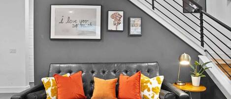 Una sala de estar moderna con un sofá de cuero negro, cojines estampados en naranja y amarillo, una cita enmarcada en la pared y una escalera con barandilla negra.